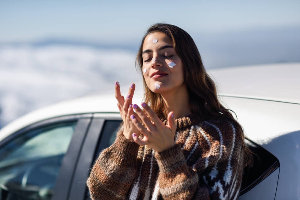 Young woman applying sunscreen on his face in snowy mountains in winter. Woman in winter clothes.