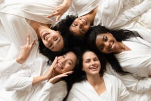 Top view five diverse women in white bathrobes on bed smile look at camera feel happy after body treatment, day spa procedures, beauty salon resort satisfied clients, bachelorette party concept